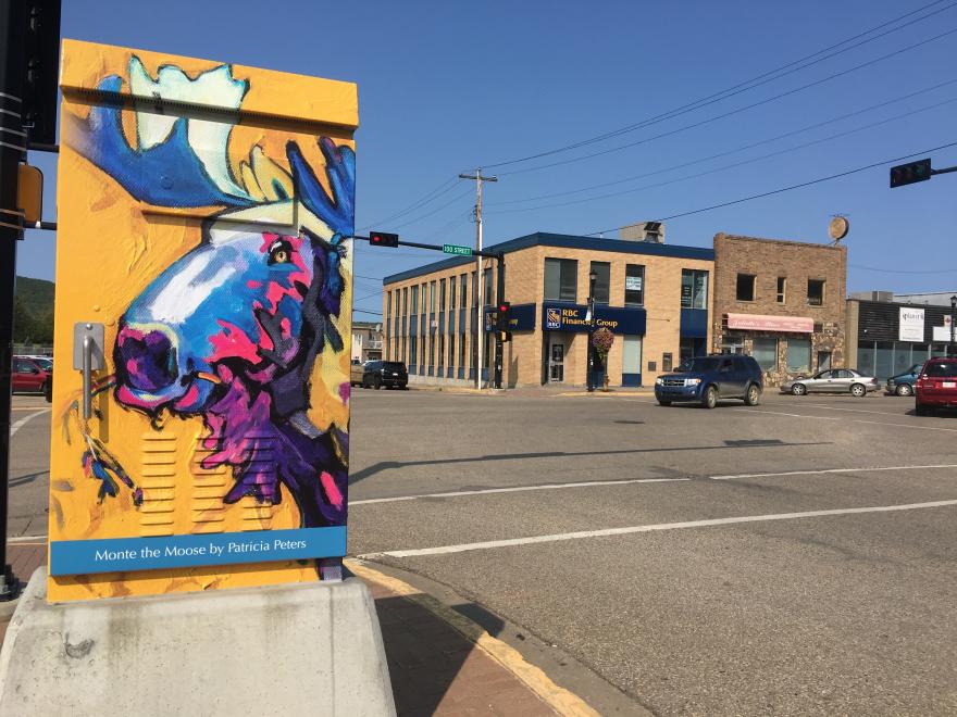 Painting of a multicolour moose head on a yellow background. The artwork is painted on an electrical box. There is a small building in the background.